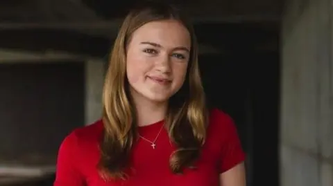 Miriam Miriam smiles at the camera. she is wearing a red top and had blonde hair
