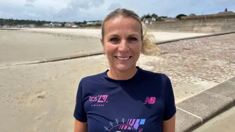 A blonde-haired woman stood on a path beside a sandy beach. 