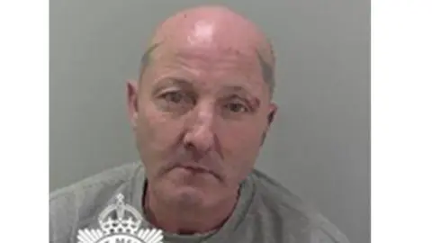 Julian Thomas in a police mugshot. He has a bald head and tattoos on both sides of his head. He is wearing a grey sweatshirt.