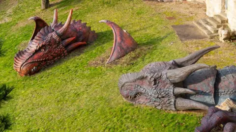 Two large, scaly dragon sculpture heads are emerging out of the grass at Lincoln Castle. One is red with black bits and has part of a wing coming out of the grass and the other is grey and you can see some of its claws in the corner of the image.