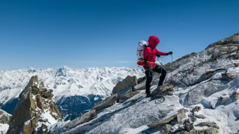 Fay Manners using ropes to reach the summit of the mountain. She is carrying equipment on her back and the sky is blue with snow covering the peaks of other mountains in the distance.