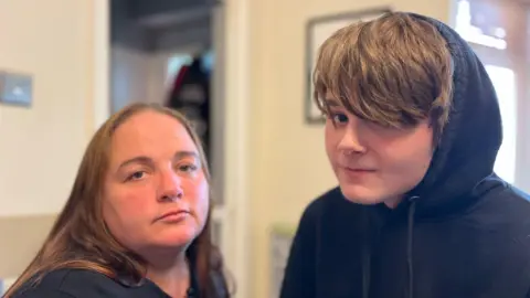 A woman who has long brown hair, wearing a black top is on the left looking towards the camera. Next to her is her 17-year-old son who has light brown hair that goes to his eyes. He is wearing a black hoodie with the hood up