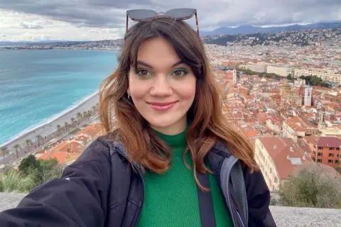 Bianca Ionici is a woman smiling for a selfie from a tower. This is a city, mountain and sea in the background