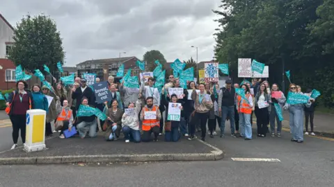 National Education Union A group of dozens of people are at a road junction, with many holding light blue banners. Some are kneeling down and others are standing up, with buildings and trees in the background.
