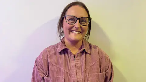 a woman in a pink shirt with brown hair tied up and wearing glasses, smiling at a camera