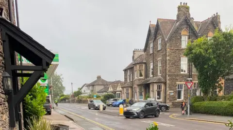 Ambleside The Wrong Way A black car drives the wrong way out of a one-way street. It is edging across the road while a hiker looks at the car. The street is in Ambleside, Lake District, and features large town-house slate buildings. 