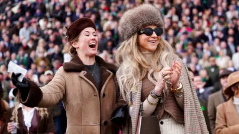 Mike Egerton/PA Two female spectators dressed in brown and cream outfits look to the right and cheer as they watch a race.