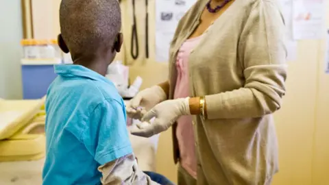 A young boy in a blue T-shirt with his back to the camera is test by HIV by a nurse a clinic that received Pepfar funding in South Africa - archive shot.