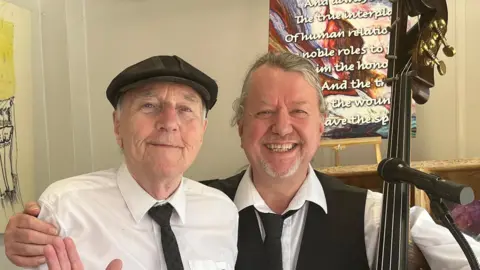 Mike Kemp in a white shirt with a tie and hat smiling. He is standing next to Geoff Tooley with grey hair and a grey beard, who is also smiling while holding an instrument. The walls are covered in colourful paintings.