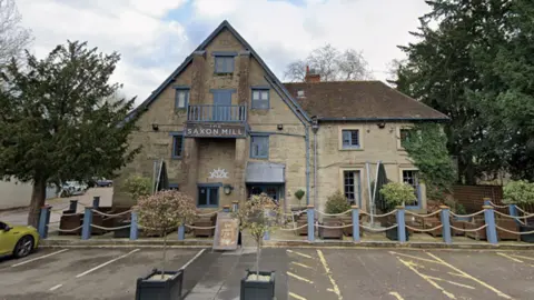 The entrance to a pub that is built from light stone. There are a five car parking spaces in front on the entrance and several bay trees. The buildings windows are painted light blue and a sign above the door reads "The Saxon Mill"