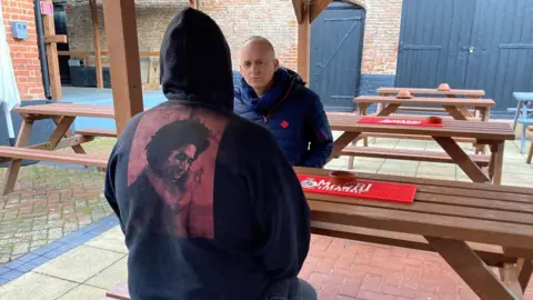 'Steve' is pictured from down  sitting connected  a picnic array  successful  a paved pub garden. He is wearing a achromatic  hooded jumper, with the hood up, and a representation   of Bob Marley printed connected  the backmost  successful  red. Opposite him is Alastair, who is wearing a bluish  padded overgarment   and scarf.