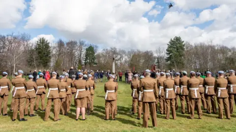 MARKSHALL Service men and women stand at a commemorative event.  Many are dressed in light brown military attire.  About 100 people in total can be seen, standing on grass at Markshall.  There is a tall stone monument and high rise trees in the background.