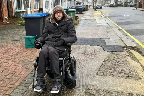 Adam Gabsi in his electric wheelchair on Scarle Road in Wembley. He is wearing glasses and dark winter clothes, including a hat with fur ear flaps. The pavements around him appear uneven