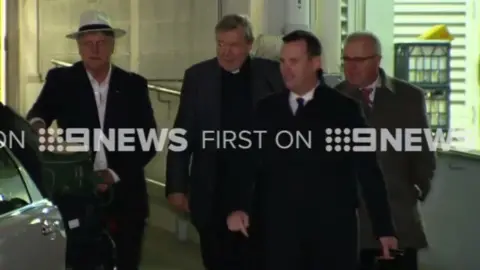 9news.com.au Cardinal Pell (second from left) arrives in Sydney, accompanied by police and private security staff