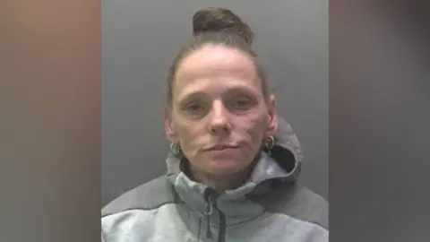 Cambridgeshire Police Adele Machin, a woman wearing a grey zip-up tracksuit with a hood. Her hair is scraped into a bun on the top of her head, she is wearing gold hoop earrings and looking directly at the camera.