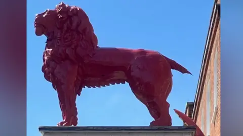 William Reid A wooden red lion sculpture stands atop a building, its mane and muscular form gleaming in the sunlight against a clear blue sky. Its tail has broken off.
