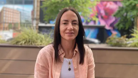 BBC A woman with dark hair looks directly at the camera. She is wearing a pale pink shirt and white vest. 