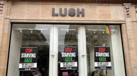BBC The shopfront of a Lush store with three posters in each window which reads: Stop starving Gaza, we are closed in solidarity. The lights inside are off