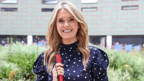 Claire Sisk/canseecantsee_ Smiling blonde woman with mid length wavy hair wearing a navy dress with white polka dots and long sleeves. She's holding a red-handled cane and standing outdoors in front of a patterned building with greenery and flowers in the background.