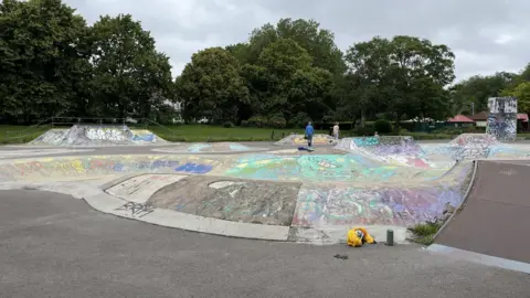BBC A skatepark is in the foreground of the picture. There are a number of ramps which are all covered in coloured graffiti. Two people can be seen at the back of the skatepark. Behind the skatepark is the green grass of the park and a row of trees.