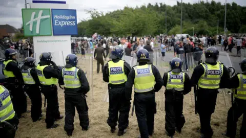 Getty Images A line of police officers wearing yellow vests, some of them are holding riot shields and batons. There is a Holiday Inn Express sign in the background. There are a large number of people standing on the street behind the police officers.