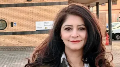 Peterborough Labour Shabina Qayyum standing in the car park of a doctor's practice. She has dark flowing hair and is wearing a green jacket and a white shirt.