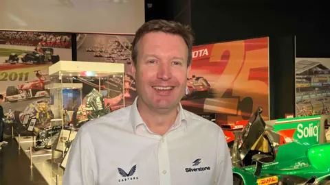 Kate Bradbrook/BBC Stuart Pringle with short brown hair smiling at the camera and wearing a white T-shirt with the Silverstone Logo. He is standing in front of display cases at an exhibition.