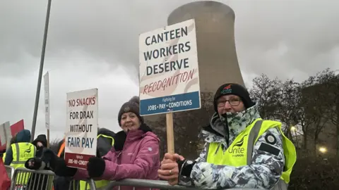 BBC Workers stand on the picket line outside Drax power station. They're are wearing high-vis and holding placards. A cooling tower can be seen in the background.