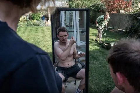 Rory Milner Man cuts his own hair in mirror as garden as other man mows the lawn