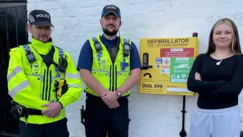 Gloucester BID Nicole on the right with two police officers standing next to a defibrillator