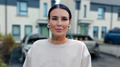 A woman, with black hair tied back, standing in front of a row of houses. She is wearing a cream coloured jumper and has white earrings. She has tattoos on her neck. 