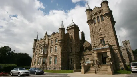 A daytime picture of Stormont Castle, a 19th-century, two-story Scottish Baronial-style mansion featuring rugged, sandstone masonry, textured walls, and ornamental turrets. Some vehicles can be seen parked at the front courtyard.