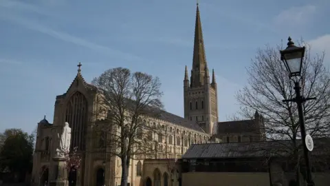 Shaun Whitmore/BBC Norwich Cathedral. A large building with a spire reaching into the sky. The picture has been taken on a sunny day with a blue sky in the background.