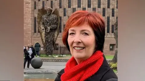 other A woman with short red hair, wearing a bright red woolly scarf and black coat looks at the camera. Behind her is a backdrop of Coventry Cathedral with the knife angel, a statue of an angel made out of knives, is behind her.