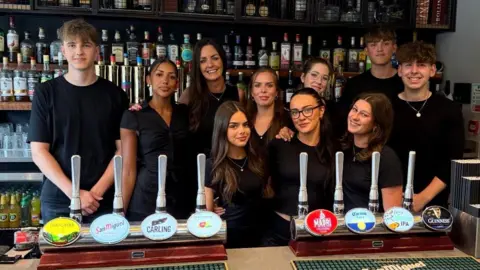 West One Image shows 10 people behind a bar, all are wearing black and smiling 