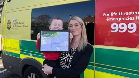 Richard, being held in his mother's arms, holding up the certificate, in front of an ambulance.