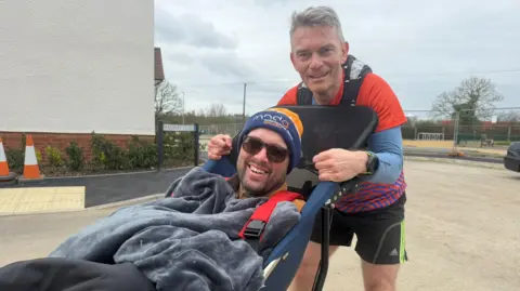 Two white men looking and smiling at the camera, one is in a wheelchair the other stands holding him.