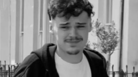 Police handout Black and white photograph of William Sullivan. He has brown curly hair and stubble. He is wearing a white t-shirt and black hoodie outside a building.