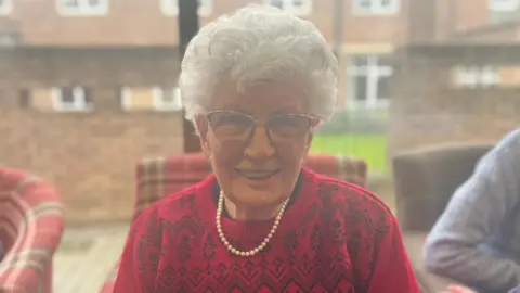 Irene Steer looking at the camera wearing glasses, a pearl necklace and a red jumper.