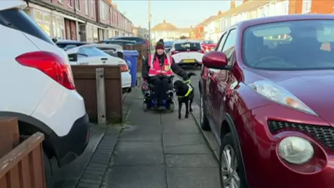 Julie in her electric wheelchair on a narrow pavement with her guide dog. A red car is parked partly on the pavement, narrowing it further. Rows of terraced housing can be seen, with more cars parked in small drivers and on the road.
