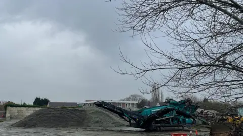 Simon Thake A large outdoor yard dominated by a high pile of a grey material similar to sand. A green and yellow digger are parked nearby 