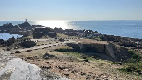 A picture of La Corbière. The lighthouse can be seen in the background to the left. There is a bunker on a hill and a car parked outside it. To the right there are houses. The sea is in the background and the sun is lighting the sea up. The sky is blue.