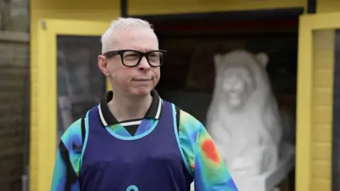 Clarke is standing in front of a small art workshop shed. It's painted yellow and a white large model of a lion is visible but out of focus. Clarke is wearing a colourful jersey and black glasses and looking to his left at someone out of frame. 