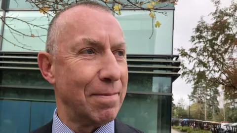 Mark Hill is standing outside a glass office. He is wearing a grey suit and blue and white shirt. He is smiling and looking off to his left.