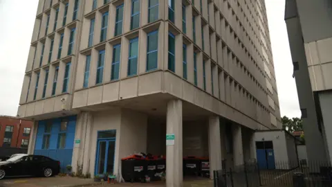 Steve Hubbard/BBC The Heights block of flats from a ground viewpoint, the building has a glass door with a blue trim and rubbish bins in a courtyard