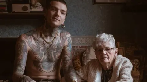 The photo Tom took, one of the winning photos in Portrait of Britain. Adam and his nan Janet sit next to each other in her house, with Janet putting both hands around Adam's left hand. He is topless, revealing tattoos of mythological creatures that cover his torso and arms. Adam has gold hoop earrings and a gold necklace. He wears brown trousers and converse-style shoes. Janet wears maroon leather shoes, brown trousers and a thick cream cardigan. She has short silver hair and wears glasses.