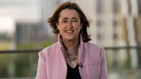 Dodds is stood in the middle of the photograph wearing a pink jacket, a necklace and black top, with large earrings. 