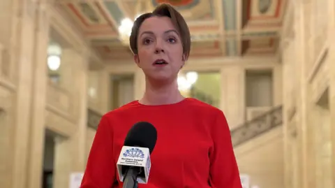 PA Media Liz Kimmins, a woman in red dress with short fair hair, facing a microphone inside Northern Ireland Assembly building