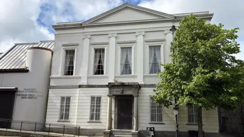 The former Newry Arts Centre - a cream-coloured two-storey building with a replica classical facade and a modern side extension. The main structure is symmetrical, with five large Georgian-style sash windows on the top floor, intersected by four decorative pilasters. The front door is protected by a metal grill and sheltered by a pillared granite portico. A disabled access ramp leads to the modern side extension which has a curved exterior wall and a steep roof.
