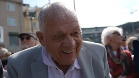 Terry Le Main. He has short grey hair and is wearing a white shirt and grey suit jacker. He is smiling. 